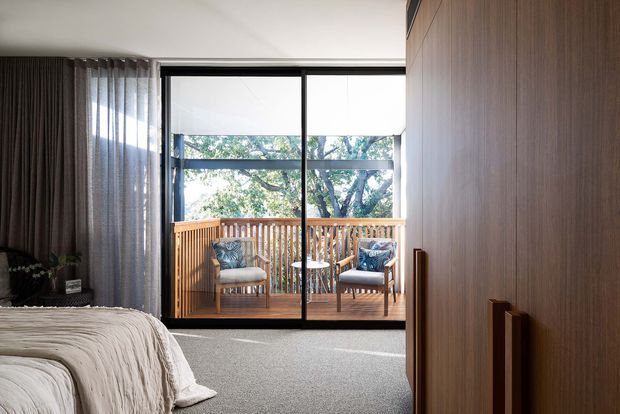 A contemporary bedroom with a partially visible bed, soft grey carpet, full-height dark curtains, and extensive timber built-in wardrobe cabinetry. Black-framed sliding glass doors open to a timber balcony with outdoor seating, bringing in abundant natural light and views of trees.