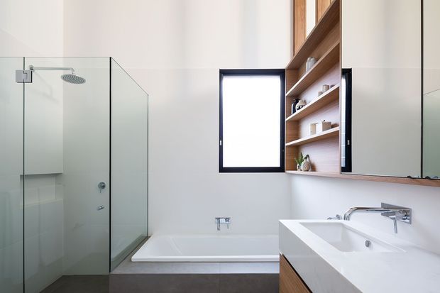 Contemporary bathroom with a frameless glass walk-in shower, freestanding-style rectangular bathtub, and a double vanity with wall-mounted chrome tapware. Warm timber open shelving and cabinetry contrast with white walls and sanitaryware, while a black-framed window adds definition. The room is bright, clean, and minimalist in character.