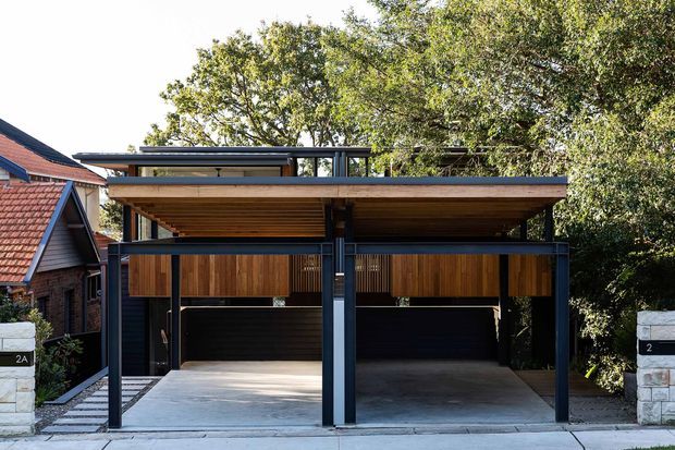 Front exterior view of a contemporary double carport attached to a residential property. The structure features a flat roof with a broad timber-lined soffit, dark steel posts and framing, vertical timber screening panels, and two open concrete parking bays. Existing brick and tiled-roof houses are visible at the sides, with mature trees behind.