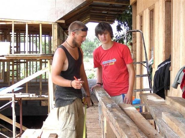 Photograph of two people working on a partially constructed timber building or elevated wooden structure. Exposed timber framing, rough-sawn boards, an open-sided upper level, and unfinished joinery dominate the scene.