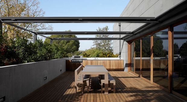 A contemporary outdoor dining terrace viewed from the deck, with timber decking, a long dining table and benches, a built-in barbecue, glass sliding doors, timber-framed glazing, and a large retractable or louvred overhead shade structure. The terrace is enclosed by smooth concrete privacy walls and timber screening, with leafy garden surroundings.