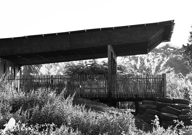Black-and-white photograph of a simple elevated outdoor shelter or pavilion with a broad, shallow sloping roof supported by timber posts. Vertical timber screening encloses the rear, while dense planting and stacked tires surround the structure. The image has a rustic, utilitarian character and strong contrast.