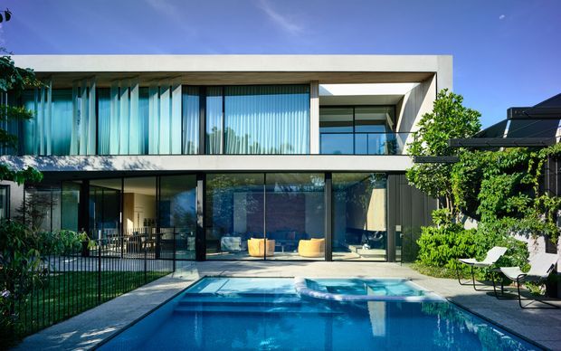 A contemporary two-storey residence with a strong rectangular, flat-roofed form, extensive floor-to-ceiling glazing, and dark window frames. The ground level opens directly onto a paved pool terrace, with a rectangular swimming pool in the foreground and landscaped planting around the property. Clean horizontal lines, recessed upper-level balconies, and a restrained palette of white concrete, dark metal framing, and glass define the design.