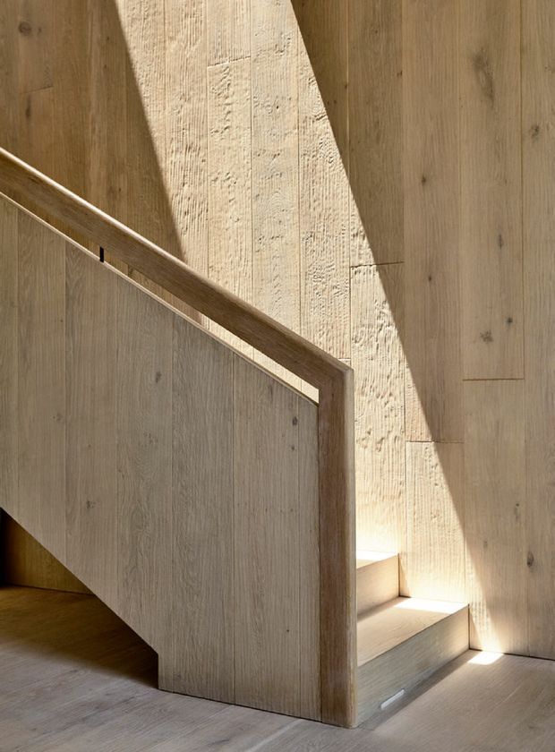 A sculptural interior stairway clad almost entirely in warm natural timber, with a solid timber balustrade and concealed stair flight. Strong sunlight creates dramatic angular shadows across the vertical wood-panelled walls and stair surfaces, highlighting the grain and tactile texture. The composition is minimalist and architectural, with no visible furnishings.