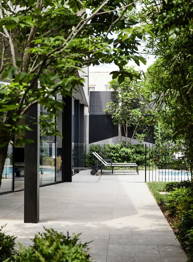A landscaped contemporary courtyard beside a modern home, with a broad pale paved terrace, floor-to-ceiling glass doors, dark structural framing and screening, lush established planting, and a partially visible swimming pool behind a black metal fence. A pair of outdoor loungers and a long low table create a relaxed poolside setting.