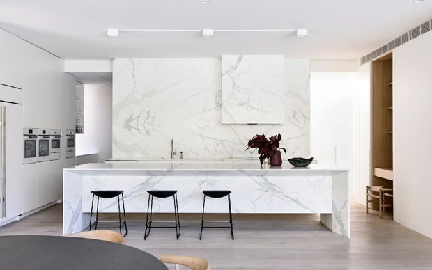 Bright, refined contemporary kitchen with a large white marble island, waterfall ends, integrated appliances, minimal white cabinetry, timber shelving, and three black bar stools. The space has light timber flooring, white walls and ceiling, concealed ventilation, and a large marble feature wall with matching slab artwork or panels.