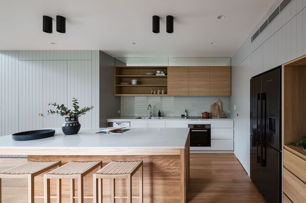 Contemporary open-plan kitchen with a large white island and integrated timber cabinetry. The space features vertical white wall panelling, warm wood finishes, a pale stone benchtop and splashback, black appliances and lighting, and woven timber bar stools. Open shelving and indoor greenery add texture while maintaining a clean, refined feel.