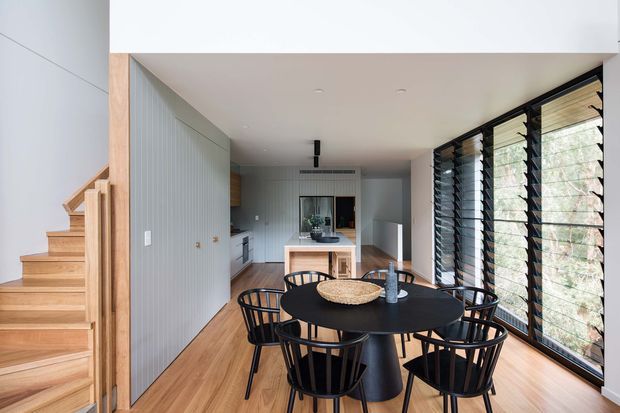 A bright, contemporary open-plan dining and kitchen area with a black round dining table and matching spindle-back chairs in the foreground. Warm timber flooring and a timber staircase with open balustrade sit to the left, while a pale vertically panelled wall and compact kitchen extend toward the rear. Full-height black-framed louvred glazing lines the right side, bringing in filtered natural light and views of vegetation. The space has a clean, refined fit-out with strong contrast between light walls, warm timber, and black furniture and window frames.