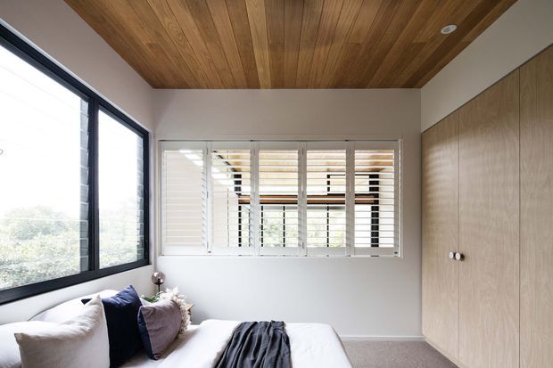 A bright, compact bedroom with a low-profile bed, layered cushions, and a dark throw. Full-height black-framed glazing on the left and plantation shutters at the rear provide natural light, while a full wall of light timber built-in wardrobes lines the right side. The ceiling is clad in warm timber boards, creating a refined contemporary interior with a calm, natural feel.