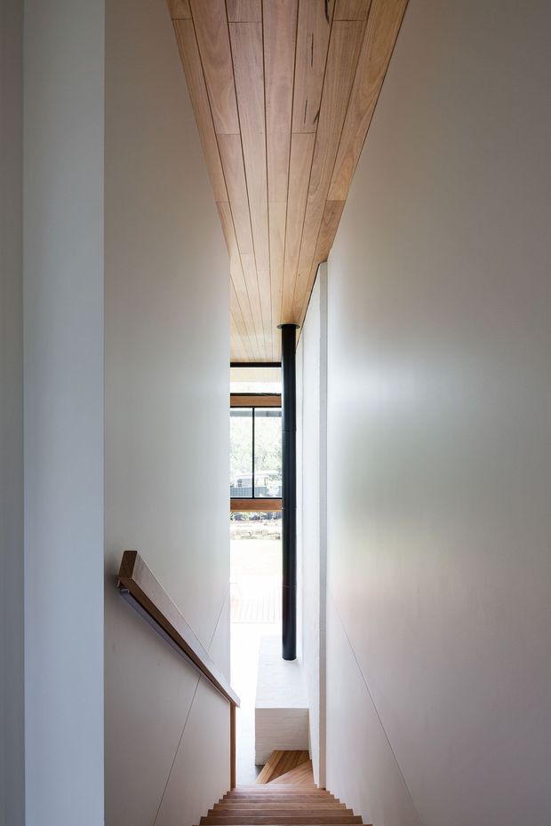 Narrow, tall interior stairwell viewed from the upper landing, with timber stair treads, a timber handrail, crisp white walls, and a warm timber-lined ceiling. A slender dark structural post and floor-to-ceiling glazing at the lower end bring in daylight and provide a view toward an outdoor area.