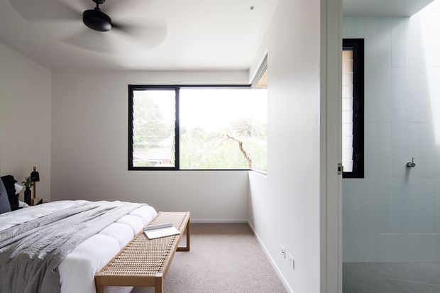 Bright contemporary bedroom with a neatly made bed, textured woven bench, and large black-framed corner window overlooking greenery. A partially visible tiled bathroom is accessed directly beside the bedroom. The room has white walls, pale carpet, a ceiling fan, and a restrained neutral palette.