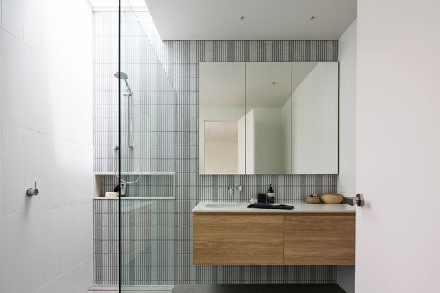 Contemporary bathroom with a walk-in glass shower, white wall tiles, vertically patterned grey tile feature wall, wall-mounted timber vanity, white basin, and mirrored cabinet. The space is clean, bright, and minimalist with black and chrome fixtures.