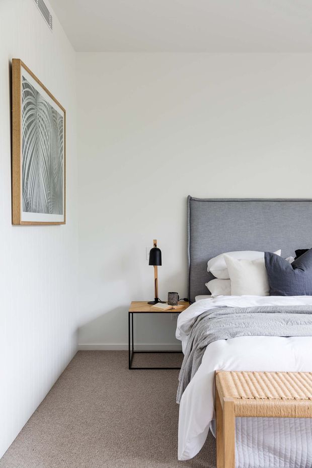 A bright, softly styled bedroom featuring a partially visible bed with a tall grey upholstered headboard, white and grey layered bedding, and dark blue accent cushions. A slim black-framed bedside table with a timber top holds a black lamp and small decor. The room has white walls, neutral carpet, a framed monochrome artwork in a timber frame, and a woven timber-framed bench at the foot of the bed.