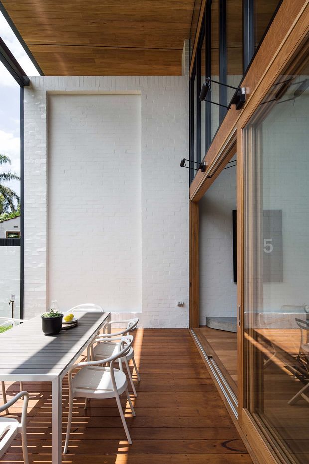 A bright contemporary courtyard-style outdoor dining terrace beside a glazed timber-clad building. The space features a long grey outdoor dining table with white chairs, warm timber decking and soffit lining, exposed white-painted brick walls, large timber-framed glass doors, and black wall-mounted exterior lights. Strong indoor-outdoor connection and abundant natural light are visible.