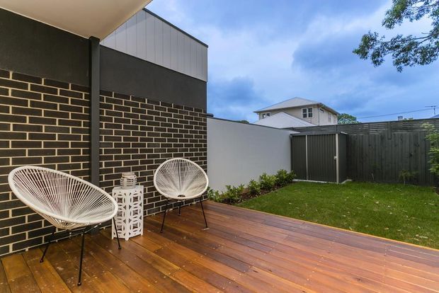 A compact landscaped courtyard with a timber deck bordered by dark brick and rendered walls. Two white woven lounge chairs and a small side table create a relaxed outdoor seating area, with a dark timber privacy fence, lawn, and neighbouring roofline visible beyond.