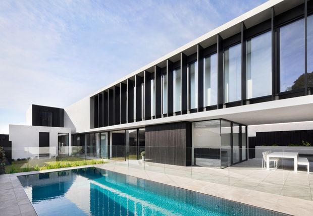 A contemporary multi-level residence with strong horizontal and geometric lines, white rendered volumes, dark vertical screening and extensive floor-to-ceiling glazing. The home opens onto a large tiled terrace and rectangular swimming pool, with a minimalist outdoor dining setting beside the building.