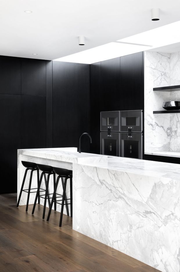 A dramatic contemporary kitchen featuring full-height matte black cabinetry, a large white marble island and matching marble splashback, integrated black appliances, a black gooseneck tap, and three black bar stools. Warm timber flooring contrasts with the monochrome cabinetry and stone, while a bright ceiling opening and minimal cylindrical downlights add a clean, refined feel.