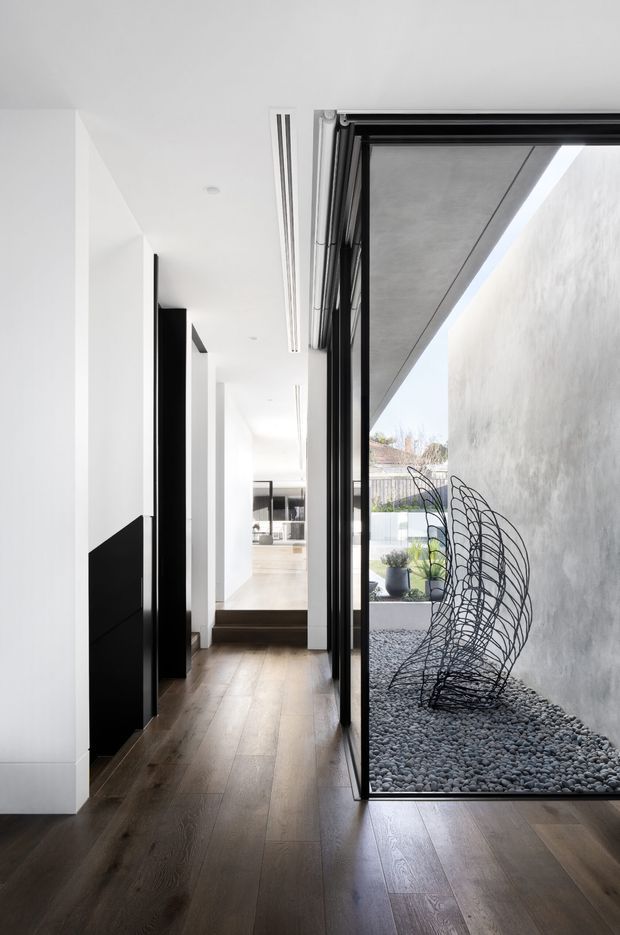 A minimalist contemporary interior hallway with dark timber flooring, white walls and ceiling, and black-framed full-height glazing. The corridor opens alongside a narrow internal courtyard or lightwell filled with smooth grey pebbles and a sculptural black wire artwork. Slim linear ceiling lights, concealed or flush doors, and strong black-and-white contrasts create a refined, gallery-like atmosphere.