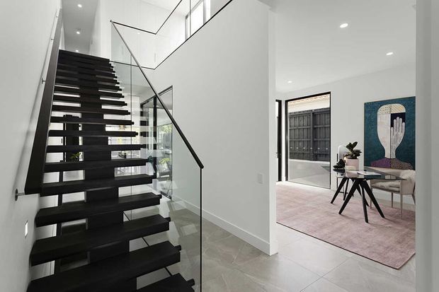 A bright contemporary interior hallway featuring a dramatic open-riser black staircase with a clear glass balustrade and slim dark handrail. White walls, recessed ceiling lights, large pale floor tiles, and an open connection to a refined sitting area with a light rug, sculptural artwork, and modern furniture are visible.