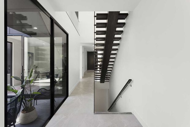 Bright contemporary hallway and stairway with pale stone or concrete-look flooring, white walls, black-framed glazed doors, and a dark open-riser staircase with slim metal handrail. A plant-filled room is visible through the glazing, while the main view focuses on the circulation space and stair void.