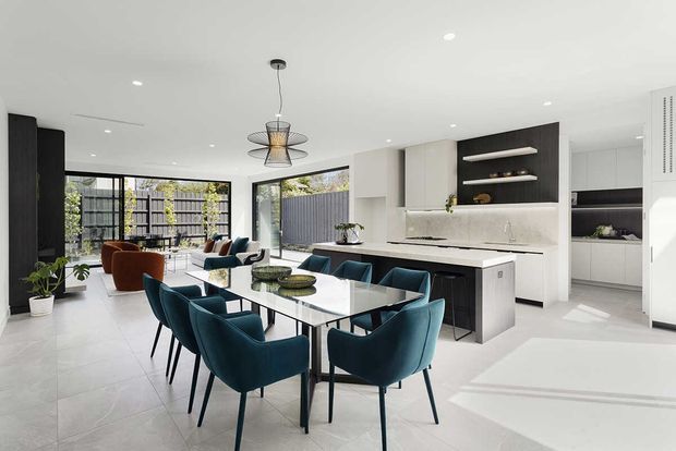 Open-plan contemporary kitchen, dining and living area with a long dining table surrounded by deep teal upholstered chairs, a white stone island with dark cabinetry, integrated shelving and a lounge seating area beyond. Large glazed doors connect to an outdoor courtyard, while recessed ceiling lights and a sculptural pendant provide illumination. The space has a bright, polished and refined feel.