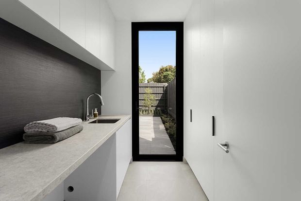 Bright, compact contemporary laundry with white built-in cabinetry, a pale stone benchtop, dark textured splashback, integrated sink and brushed metal tapware. A full-height black-framed glazed door opens to an outdoor path, while folded towels sit on the counter.