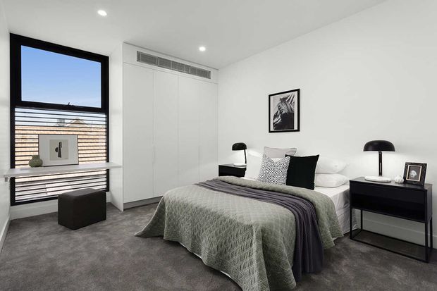 A contemporary bedroom with a low-profile upholstered bed dressed in a textured sage-green quilt and dark grey throw, black and white cushions, and matching black bedside tables and lamps. The room has soft grey carpet, white built-in wardrobes, recessed ceiling lights, a large black-framed window with horizontal blinds, and restrained monochrome artwork.