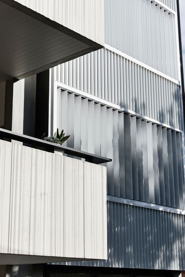 Close-up architectural exterior view of a contemporary multi-level building. The facade is composed of pale ribbed vertical metal cladding and projecting angled fins, with dark structural recesses, concrete balcony or wall planes, and a small planter on a balcony edge. The image emphasizes layered facade geometry, texture, and strong vertical lines.