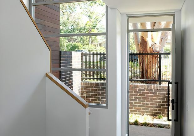 Bright contemporary entry stairway with white walls, a timber-trimmed staircase, and floor-to-ceiling glazing beside a glazed door. The view opens to a small brick-paved courtyard or front garden enclosed by a brick wall and black metal fencing, with mature trees beyond.