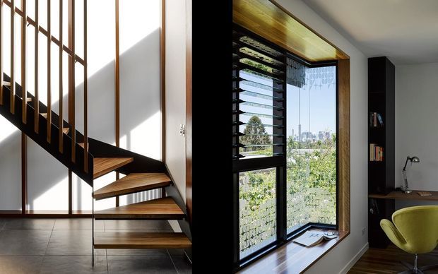 Split view of a bright contemporary interior featuring a sculptural open-riser timber staircase with dark metal balustrade on the left, and a compact home office on the right with a built-in dark shelving unit, desk, lime-green chair, and large black-framed louvred glazing overlooking greenery and a city skyline.