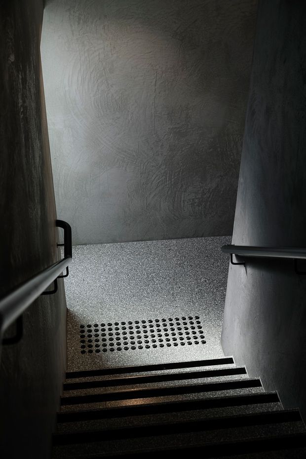 A narrow, dimly lit stairway descending to a landing. The space has dark textured plaster walls, black metal handrails on both sides, and speckled terrazzo-like flooring with a rectangular tactile dot surface at the bottom landing. Strong side lighting creates a moody, minimalist architectural atmosphere.