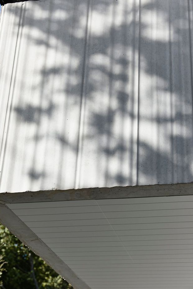 Close-up upward view of a building exterior roof edge and soffit. The upper portion shows light grey corrugated metal roofing with vertical ribs and strong dappled tree shadows. Below is a white horizontally lined panelled soffit beneath a rough concrete or rendered fascia, with blurred greenery visible at the edge. The image is architectural and detail-focused, with no interior space shown.