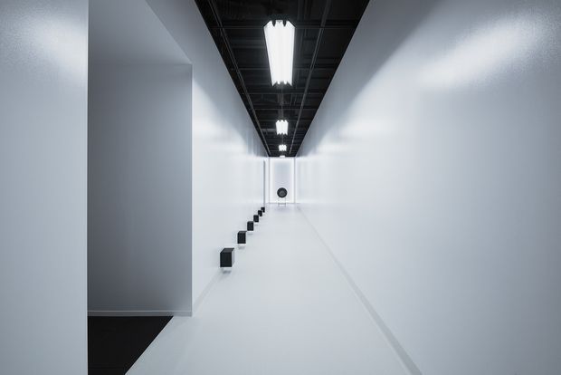 A stark, minimalist interior corridor or gallery-like passage with white walls and floor, a dark exposed ceiling grid, and a central line of bright fluorescent strip lights. Small black geometric wall or floor-mounted elements lead toward a distant circular feature, creating strong linear perspective and high contrast.