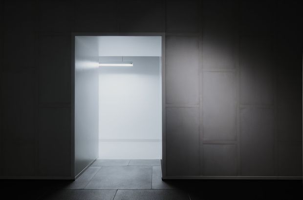 A dark, minimalist interior view looking through a wide doorway into a bright, empty room. The foreground walls and door surround are clad in dark charcoal panels with subtle vertical and rectangular detailing, while the rear space has pale smooth walls, a narrow horizontal wall light, and large grey floor tiles. The strong contrast between the shadowed foreground and illuminated interior creates a dramatic, gallery-like atmosphere.