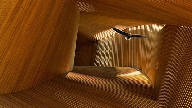 A dramatic architectural interior viewed upward through a tall, sculptural timber-lined void. Closely spaced vertical and angled wood slats wrap the walls and ceiling, creating strong rhythmic lines, warm amber tones, and patterned sunlight. A dark ceiling fan is suspended near the upper opening.