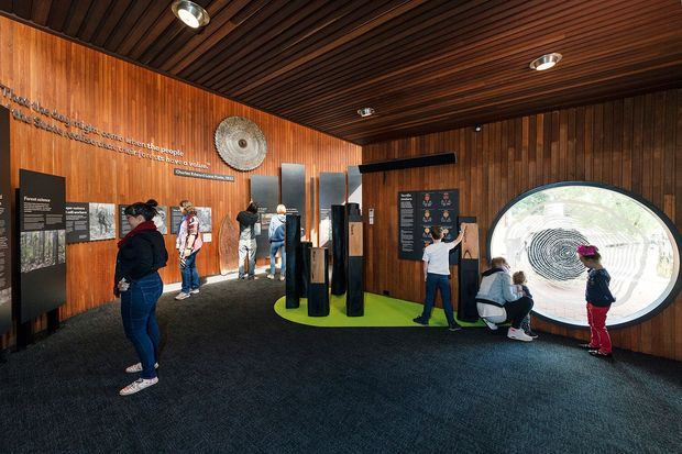 Interior view of a compact exhibition or visitor-centre gallery with dark timber wall panelling and a slatted timber ceiling. Interpretive display boards, sculptural black columns, a large circular viewing or interpretive window, and several visitors fill the space. The room has dark carpet, recessed downlights, and a warm, museum-like atmosphere.