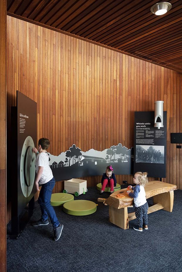 A small children's interactive exhibit or play area with warm timber-lined walls and a dark timber slatted ceiling. The space includes interpretive display panels, a wall-mounted interactive feature, low green seating, children's play equipment, and a rustic solid-wood picnic-style table. Dark carpet covers the floor, and a round ceiling light provides illumination.