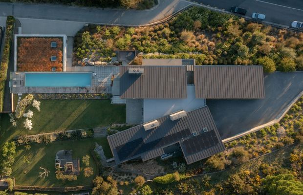 Aerial, top-down view of a landscaped residential property with several connected buildings featuring long pitched roofs, a rectangular swimming pool, paved outdoor areas, lawns, dense ornamental planting, and surrounding driveways and roads.