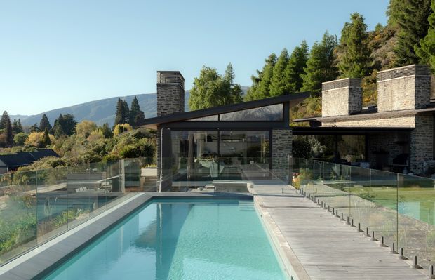 Contemporary hillside residence viewed from the pool terrace, featuring dark stone masonry walls, expansive floor-to-ceiling glazing, low-slung roof forms, a timber pool deck, and clear glass balustrades. The long rectangular swimming pool and landscaped setting dominate the foreground, with wooded hills visible beyond.