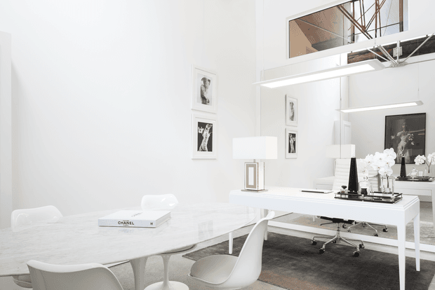 Bright, high-key contemporary dining and living workspace with a white marble dining table and sculptural white chairs in the foreground. A white desk or console sits against a large mirrored wall, with modern office chairs, framed monochrome artwork, orchids, and linear pendant lighting. The room has a lofty feel with exposed ceiling services and a warm timber-toned upper opening visible in the mirror.