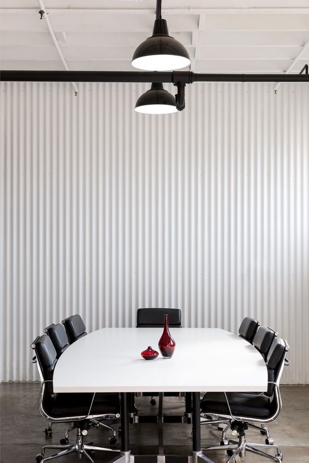 A modern industrial dining or conference room with a large white rectangular table surrounded by black leather office-style chairs. The space features a white corrugated metal wall, exposed white ceiling structure, black exposed piping, and black dome pendant lights. Two red glass vases provide a small accent on the table.
