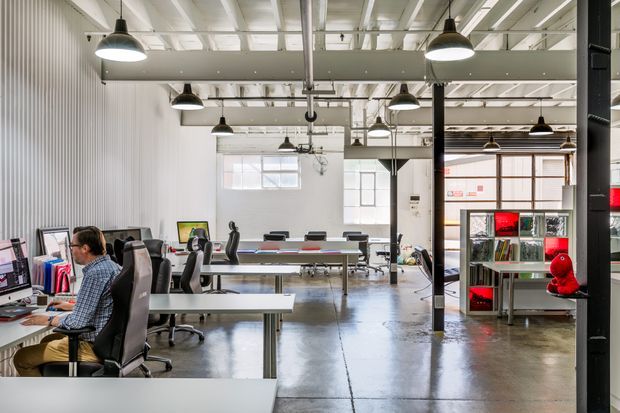 A bright open-plan creative office in an industrial-style building, with long shared workbenches, computer workstations, office chairs, exposed services, suspended black pendant lights, polished concrete flooring, corrugated metal wall cladding, and structural steel columns. A shelving unit with books and red display accents is visible at the right.