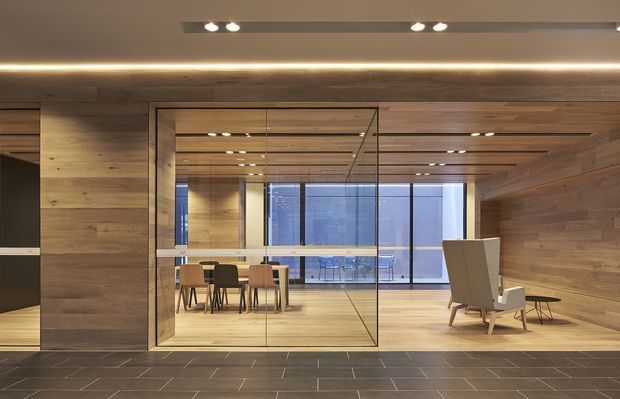 A contemporary interior clad extensively in warm horizontal timber panelling, with a glass-enclosed dining area centred in the frame. The dining space contains a table and chairs and opens visually toward floor-to-ceiling glazing. A timber-lined lounge area with an armchair and side tables is visible to the right. Dark rectangular floor tiles, recessed ceiling spotlights, and concealed linear lighting create a refined, warm commercial or hospitality-like setting.