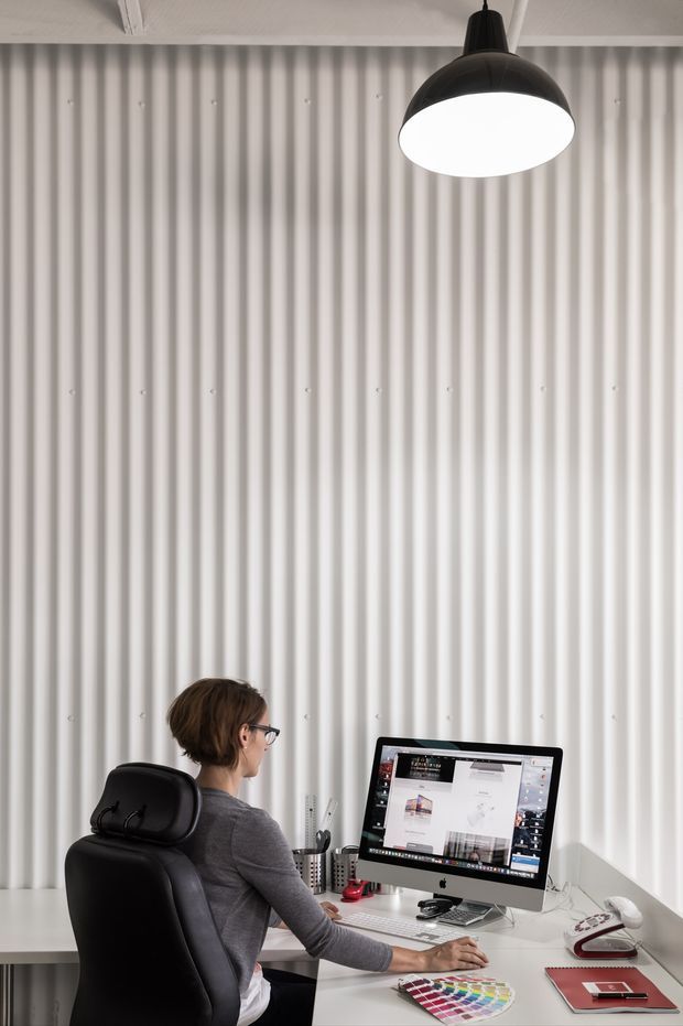 A compact, minimalist home office with a person working at a white desk. The room features a tall white corrugated wall panel backdrop, a large black pendant light, a black ergonomic office chair, and a desktop computer with office accessories. The palette is predominantly white, grey, and black with small red and multicoloured accents.