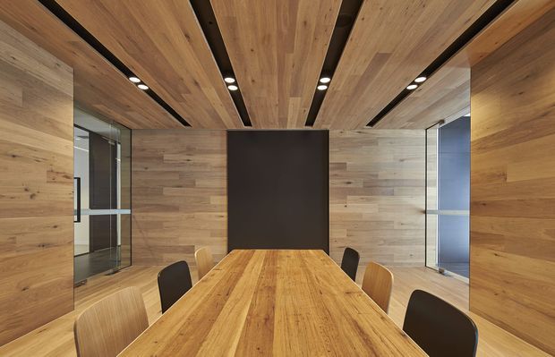 A contemporary meeting or dining room dominated by warm timber wall, ceiling, and floor finishes. A long solid-wood table is surrounded by mixed timber and dark upholstered chairs. Linear black ceiling recesses contain recessed spotlights, while glazed openings on both sides reflect the room and reveal adjacent spaces. The design is clean, highly coordinated, and minimalist with strong linear detailing.