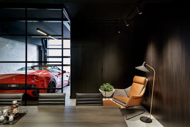 Dark, sophisticated modern interior resembling a private lounge or office, with dark timber wall panelling, exposed black ceiling services, a large timber table, leather seating, a sculptural floor lamp, and a red sports car displayed behind black-framed industrial glazing.