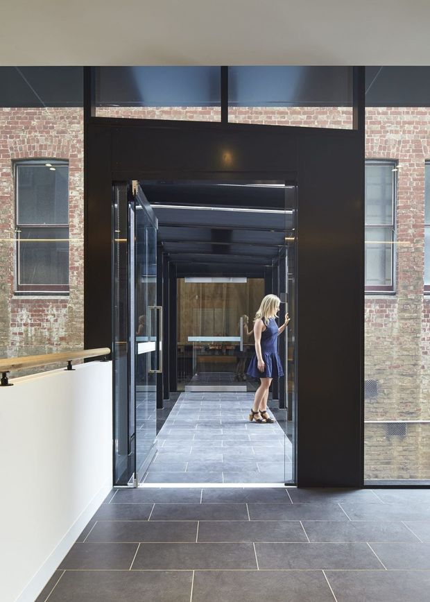 A contemporary commercial-style entry or passageway with dark charcoal tiled flooring, black metal-framed glazing and doors, and exposed red brick walls. The view looks through a series of glazed portals into a reflective corridor, with a woman standing in the distance. A pale curved balustrade with a timber handrail is visible on the left.