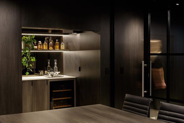 A dark, sophisticated dining area with a built-in bar and wine refrigerator. The bar features dark wood cabinetry, a light stone benchtop, illuminated open shelving with bottles and glassware, and integrated planting. A large timber dining table and black upholstered chairs occupy the foreground, while a black-framed glazed door or partition is visible to the right. The space is low-lit and highly refined with concealed shelf lighting.