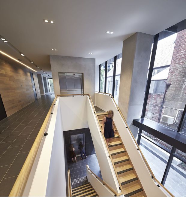 A bright, modern multi-level interior circulation space with a central open stairwell and multiple flights of stairs descending around a void. The space features large floor-to-ceiling windows, exposed concrete-toned walls and columns, dark tiled flooring, timber wall panelling, sleek metal-and-glass balustrades, recessed ceiling lighting, and elevator doors along the corridor.