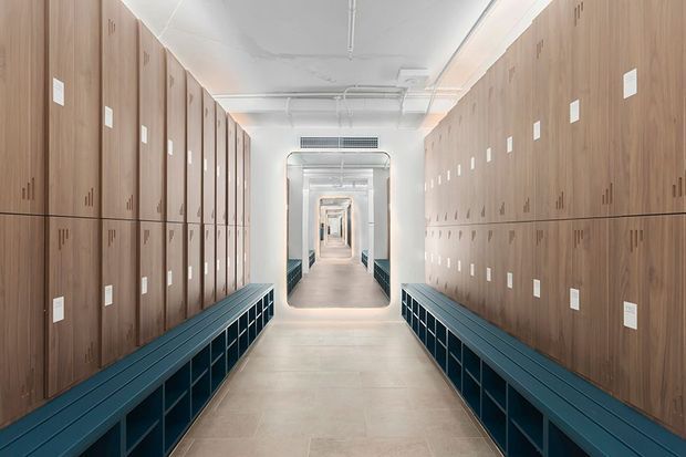 A long, symmetrical institutional corridor lined with full-height timber locker cabinetry on both sides. Dark teal built-in bench seating with open cubby storage runs along the base of the lockers, while a pale tiled floor, white ceiling with exposed services, and repeated doorways create a strong perspective toward the far end.